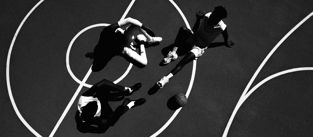 Three basketball players sit on a court in bright sunlight, seen from above. Their shadows are sharp, and a basketball rests near one player. The court’s white lines are clearly visible.