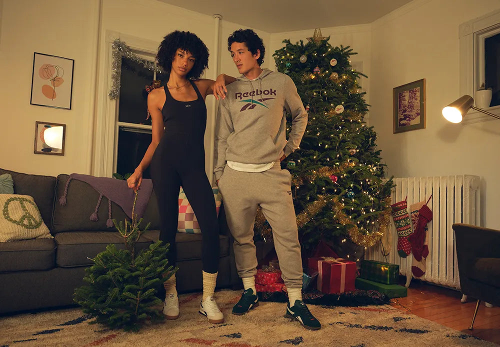 Two people pose in a cozy living room decorated for Christmas. The woman holds a small tree, and the man wears a Reebok hoodie. There’s a decorated Christmas tree, wrapped gifts, and stockings hanging by a radiator in the background.