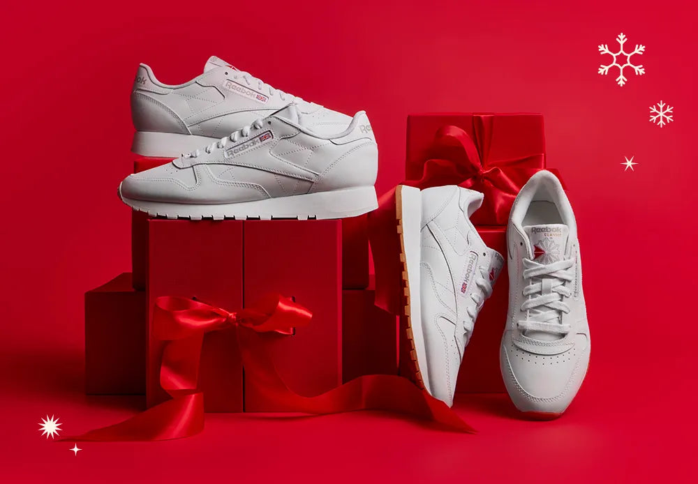 Three pairs of white Reebok sneakers displayed on and around red gift boxes with red ribbons, set against a red background with white snowflake and star illustrations, creating a festive holiday theme.