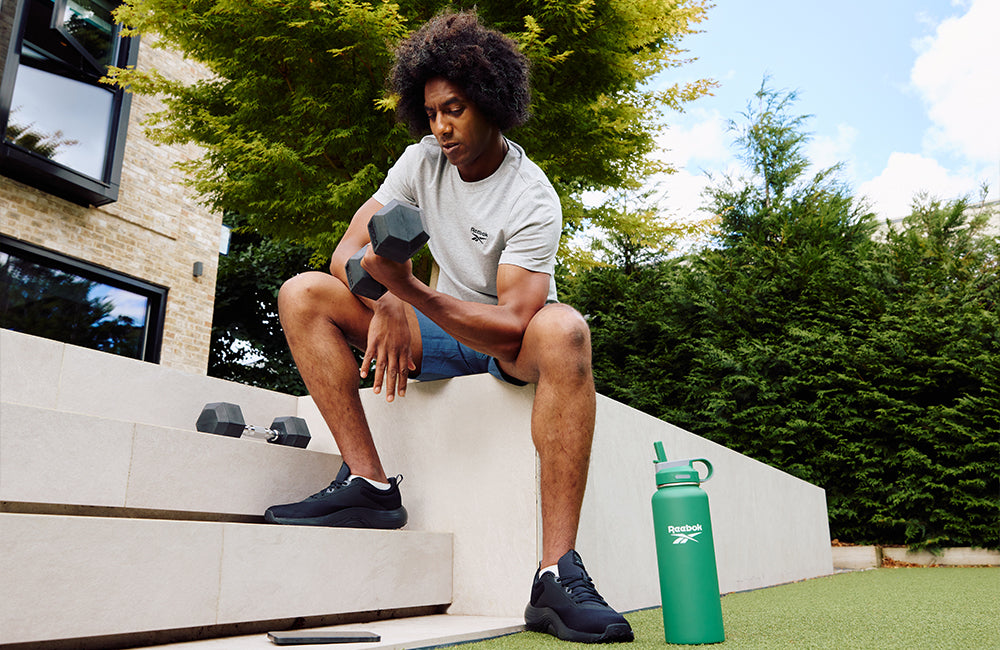 A man sits outdoors on white steps, lifting a dumbbell with one arm. He wears a grey T-shirt, blue shorts, and black sneakers. A green water bottle and more dumbbells are beside him, with trees in the background.