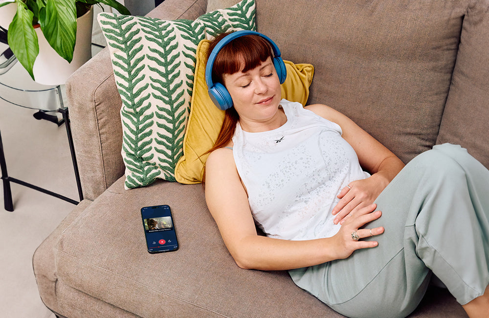 A woman with red hair relaxes on a sofa with eyes closed, wearing blue headphones. A smartphone showing a video call screen rests beside her. She lies on green and yellow cushions.