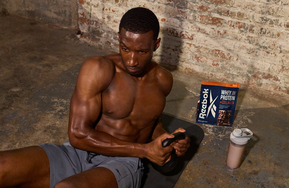A muscular man in gym shorts sits on the floor doing a core workout with a kettlebell. Beside him are a protein powder bag labeled Reebok Whey Protein + Collagen and a shaker bottle with a drink.