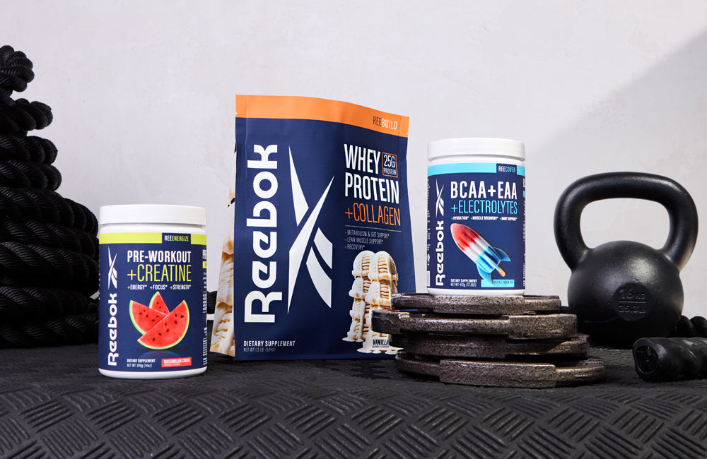 Three Reebok supplement containers—Pre-Workout with Creatine, Whey Protein + Collagen, and BCAA + EAA with Electrolytes—are displayed with black gym weights, a kettlebell, and coiled rope on a gym floor.