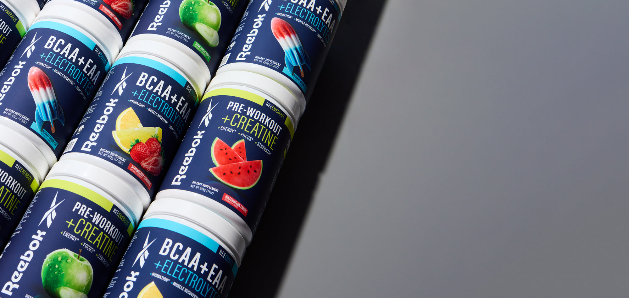 Several Reebok supplement containers with colorful fruit flavors, such as watermelon, popsicle, and apple, are arranged diagonally on the left against a reflective gray background.