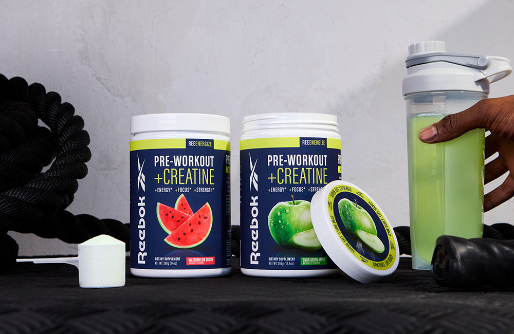 Two Reebok tubs of pre-workout and creatine powder, one watermelon flavor and one green apple flavor, sit on a gym mat. A shaker bottle with green drink and a hand, scoop, and black rope are also visible.