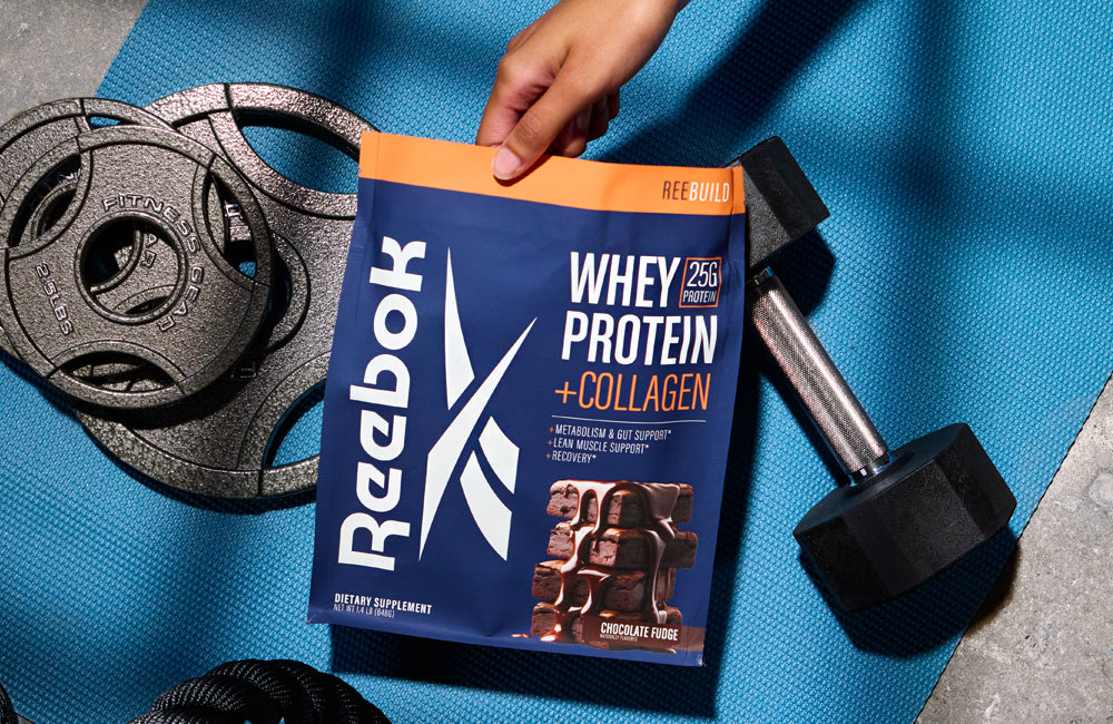 A hand holds a Reebok Whey Protein + Collagen powder pouch on a blue exercise mat, surrounded by a dumbbell, weight plates, and a coiled rope.