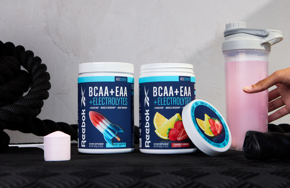 Two Reebok BCAA+EAA +Electrolytes supplement containers, one closed and one open, sit on a black surface with a pink drink in a shaker bottle nearby and a hand reaching for it. Black gym ropes are in the background.