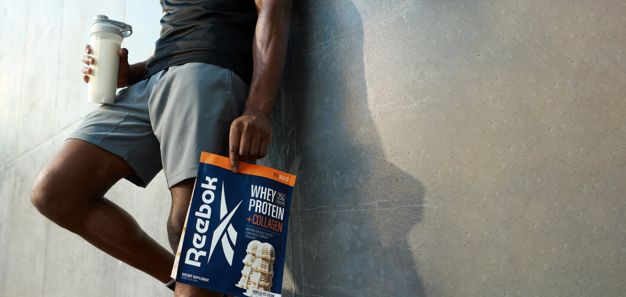 A person in athletic wear leans against a wall, holding a shaker bottle in one hand and a Reebok Whey Protein + Collagen bag in the other. Only the lower half of their body is visible.