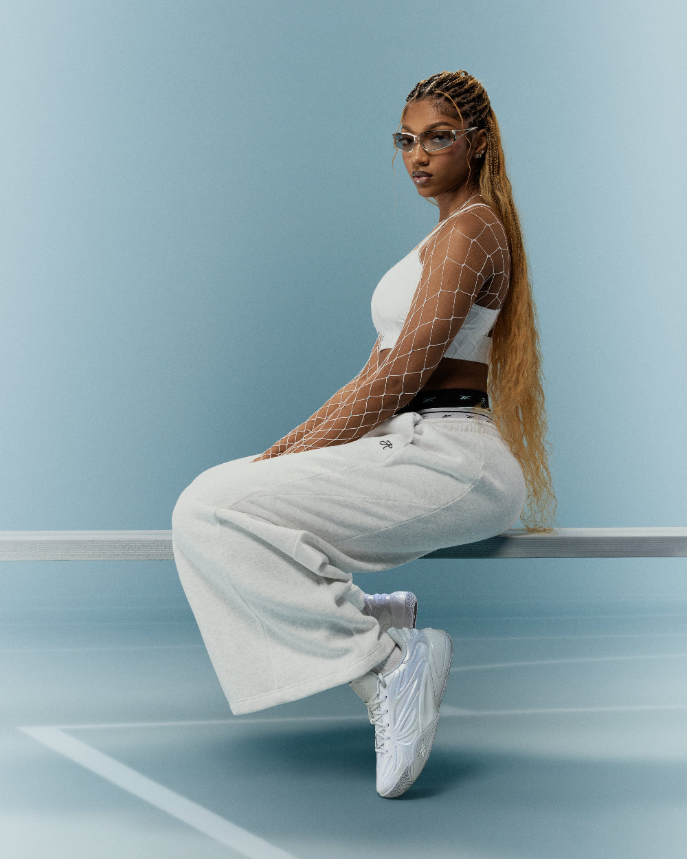 Angel Reese sitting on a bench wearing a white top, fishnet sleeves, and light gray pants against a blue background