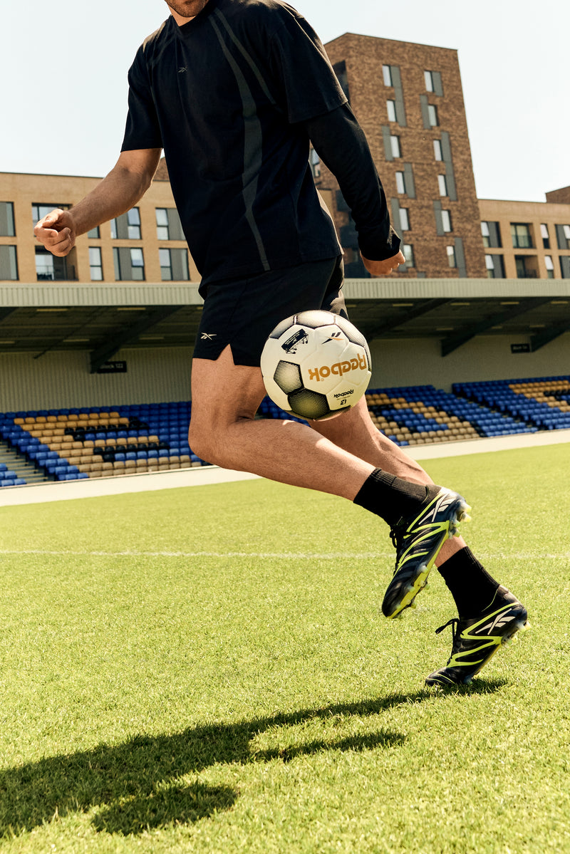 Wearing Reebok Sidewinder 26 Elite FG Cleats, Serbian soccer player Dušan Vlahović in black sportswear juggles a ball on firm grass, with empty blue and yellow seats and apartment buildings visible in the background.