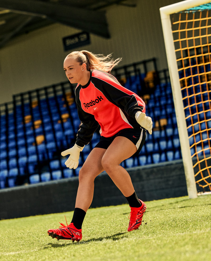 A female soccer goalkeeper in a red Reebok jersey and gloves stands focused in front of the goal on a sunny day, wearing Reebok Sidewinder 26 Elite Firm Ground Cleats planted firmly on the well-maintained grass, with empty blue and yellow stadium seats in the background.