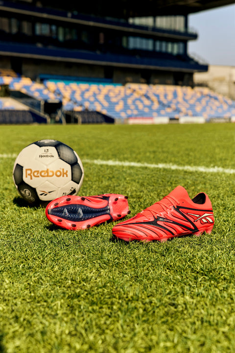 A pair of red Reebok Sidewinder 26 Elite Firm Ground Cleats and a black-and-white Reebok soccer ball, perfect for ball control, are displayed on green grass in a stadium with blue and yellow seats in the background.