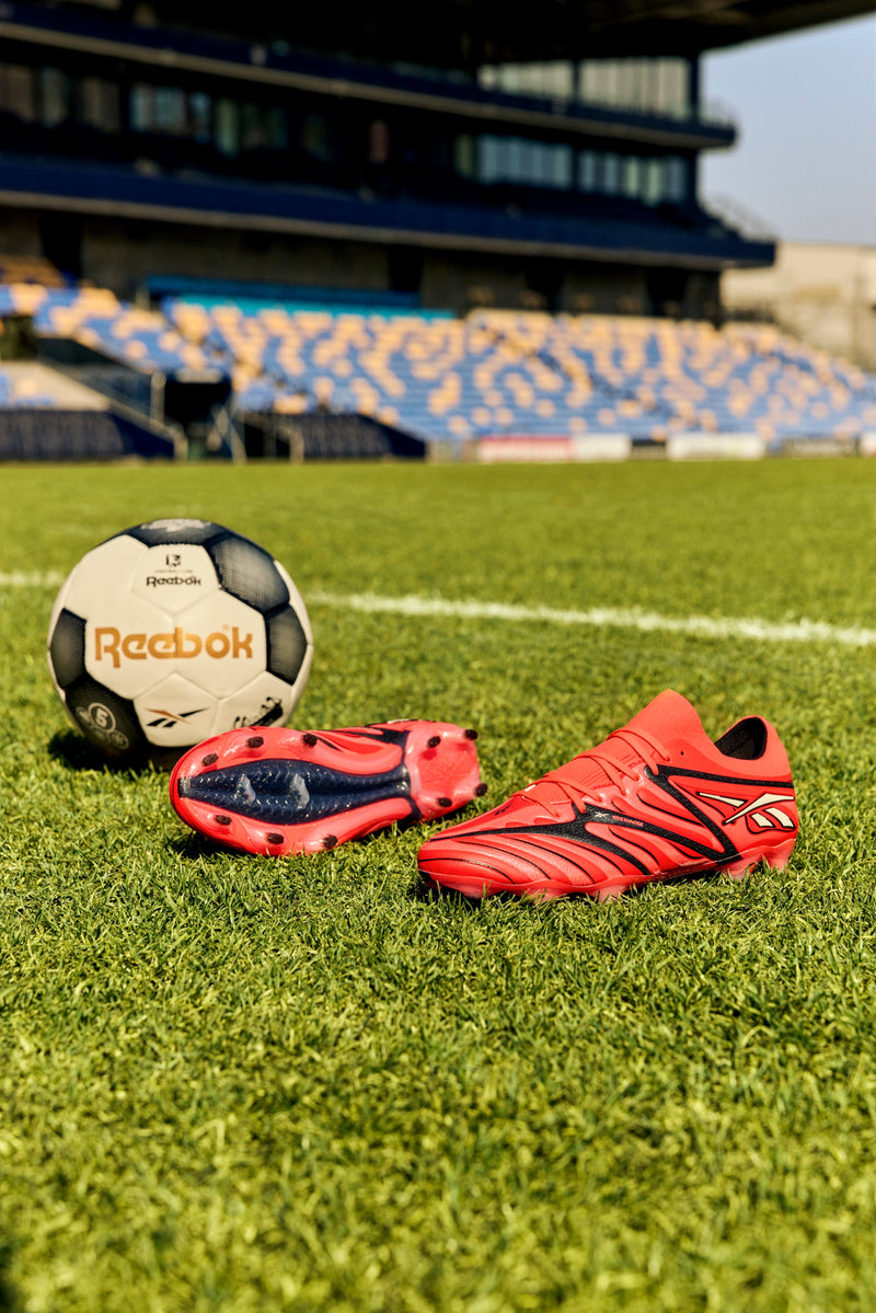 A pair of red Reebok Sidewinder 26 Elite Firm Ground Cleats and a black-and-white Reebok soccer ball, perfect for ball control, are displayed on green grass in a stadium with blue and yellow seats in the background.