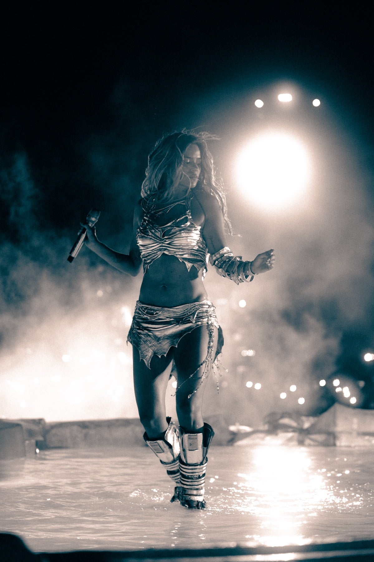 A performer in a metallic, tattered outfit dances on a wet stage at night, holding a microphone, with bright lights and mist creating a dramatic atmosphere.