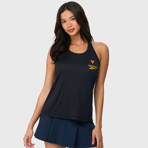 A woman with long dark hair wears the Reebok x GLDN PNT Women’s Tank Top featuring a yellow logo and a navy pleated skirt, standing against a plain light background.