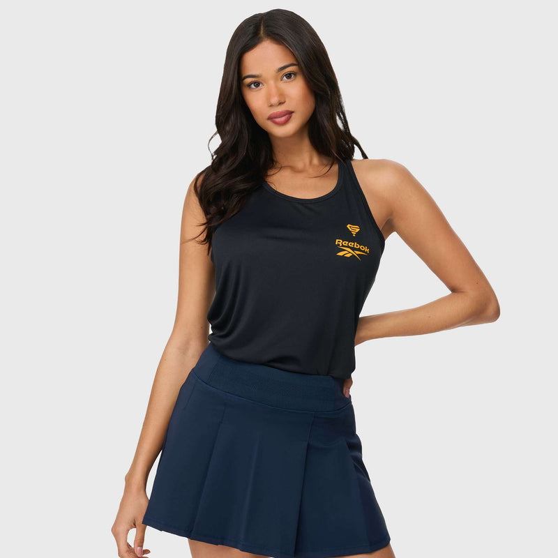 A woman with long dark hair wears a sleeveless black Reebok x GLDN PNT Women’s Tank Top with a yellow logo and a navy tennis skirt, standing with one hand on her hip against a plain light background.