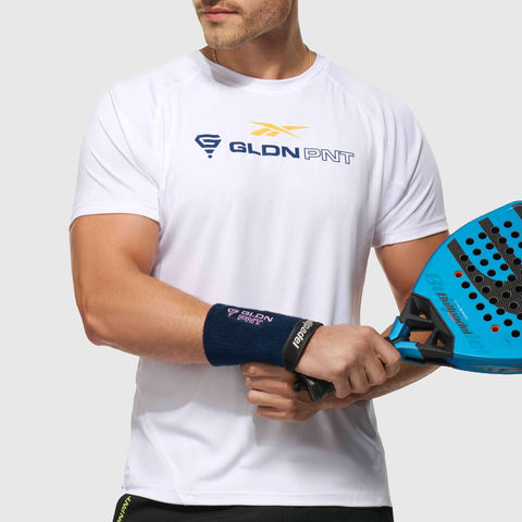 A man wearing a white Reebok x GLDN PNT Tech Tee and a wristband holds a blue padel racket, standing against a plain light background.
