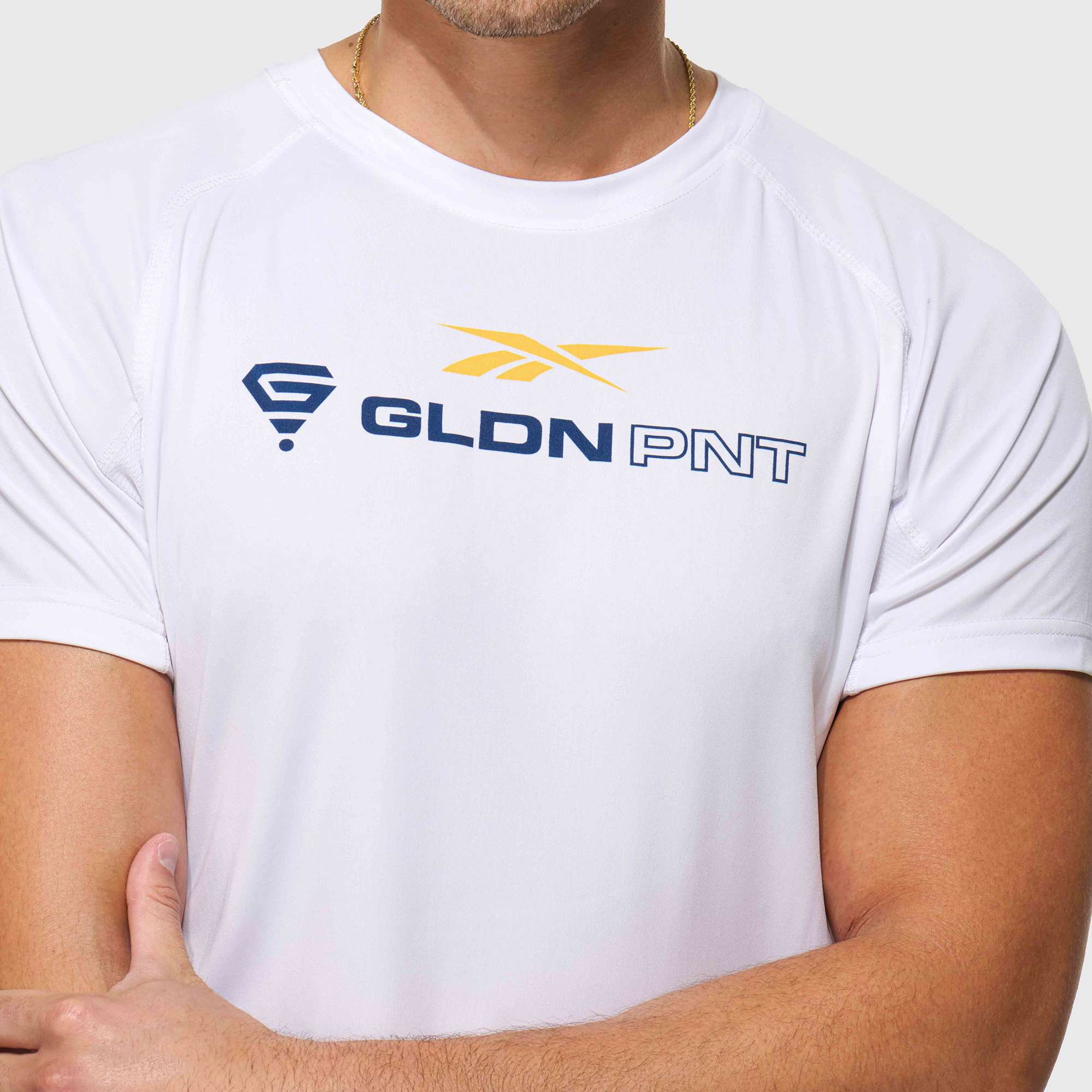 A man wears a Reebok x GLDN PNT Tech Tee featuring a yellow and blue logo on the chest. Only his upper torso and arms are shown, with his face not visible. The lightweight tee is crafted from Reebok’s moisture-wicking fabric.