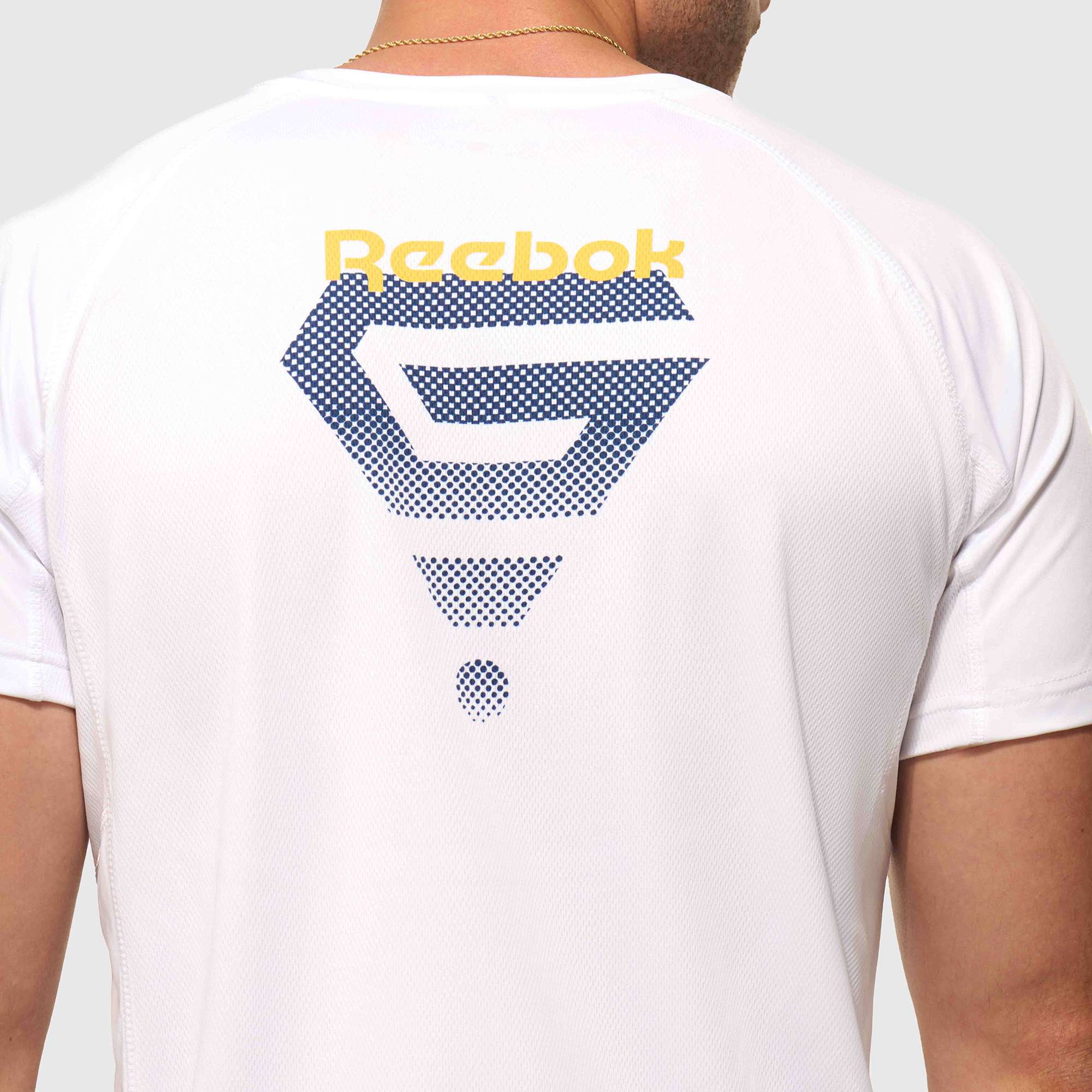 A man, seen from the back, wears a Reebok x GLDN PNT Tech Tee with a large blue geometric graphic and a yellow Reebok logo on the upper back.