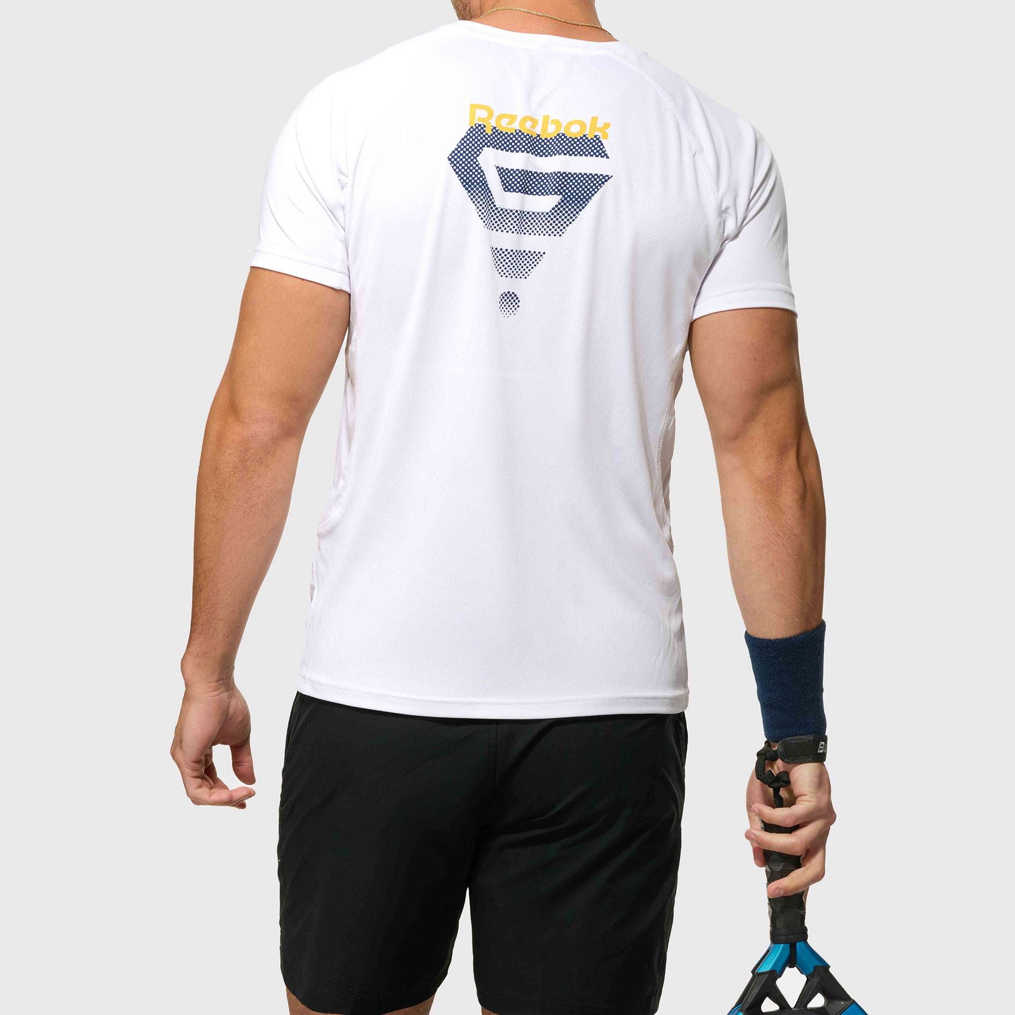 A man faces away, wearing a white Reebok x GLDN PNT Tech Tee with a blue and yellow geometric logo on the back, black shorts, navy wristband, and holding a blue and black tennis racket by his side.