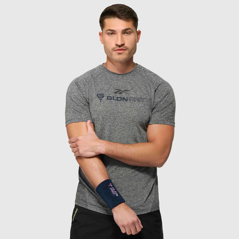A man wearing a grey Reebok x GLDN PNT Tech Tee and black shorts stands against a plain background, holding his left arm with his right hand. He has a navy wrist support on his left wrist and a neutral expression.