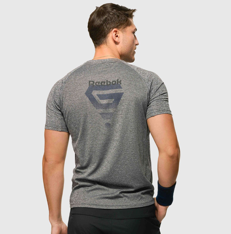A man stands facing away against a plain light background, wearing a gray Reebok x GLDN PNT Tech Tee with a large back logo and a dark wristband on his right wrist.