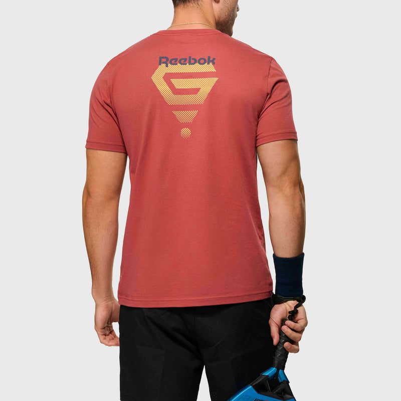 A man stands with his back turned, wearing the Reebok x GLDN PNT Jersey Tee in red featuring a yellow geometric print and Reebok branding on the back, paired with black shorts and a wristband, holding a blue paddle in his right hand.