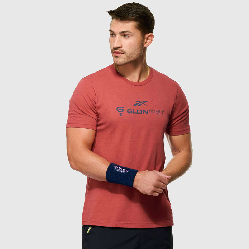 A man wearing a Reebok x GLDN PNT Jersey Tee in red with a front logo stands against a light gray background, looking left. He sports a navy wristband on his right wrist and holds his left wrist.