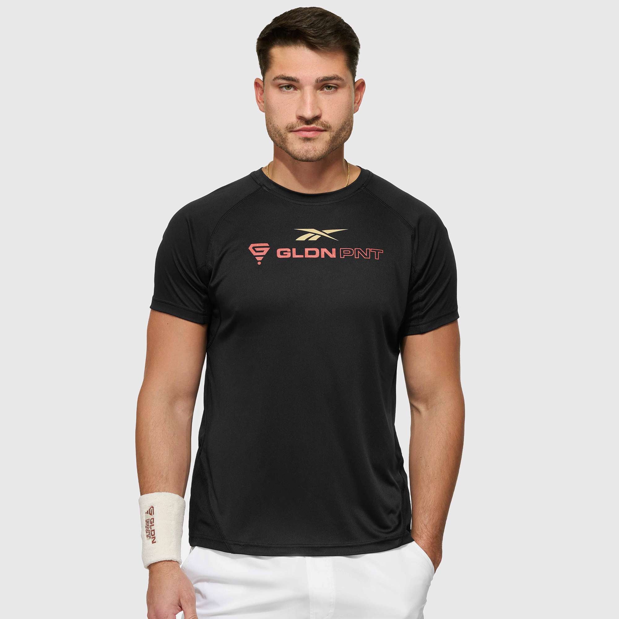 A man with short brown hair and a trimmed beard wears a black Reebok x GLDN PNT Tech Tee with gold and red lettering, white shorts, and a white wristband on his left wrist, standing against a light gray background.