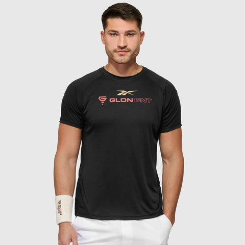 A man with short brown hair and a trimmed beard wears a black Reebok x GLDN PNT Tech Tee with gold and red lettering, white shorts, and a white wristband on his left wrist, standing against a light gray background.