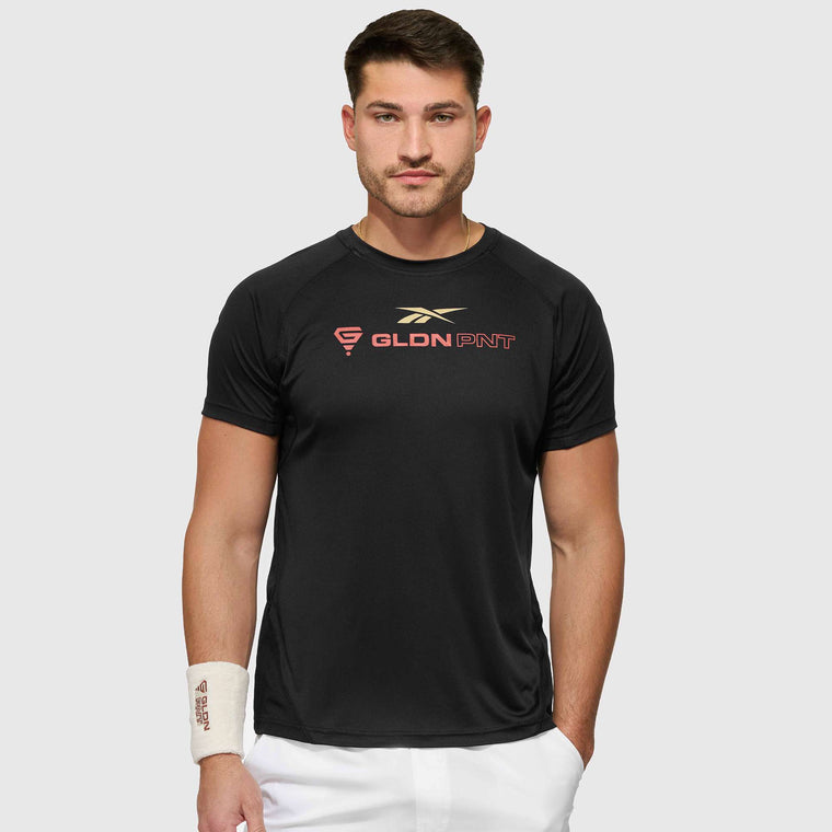 A man with short brown hair and a trimmed beard wears a black Reebok x GLDN PNT Tech Tee with gold and red lettering, white shorts, and a white wristband on his left wrist, standing against a light gray background.
