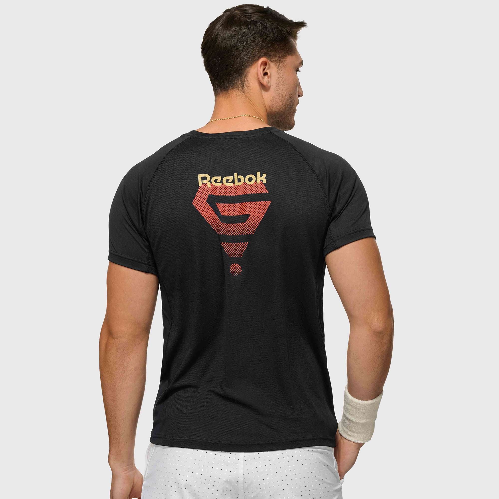 A man wearing a black Reebok x GLDN PNT Tech Tee with a large red and yellow logo on the back stands facing away, paired with white shorts and a beige wristband on his left wrist.