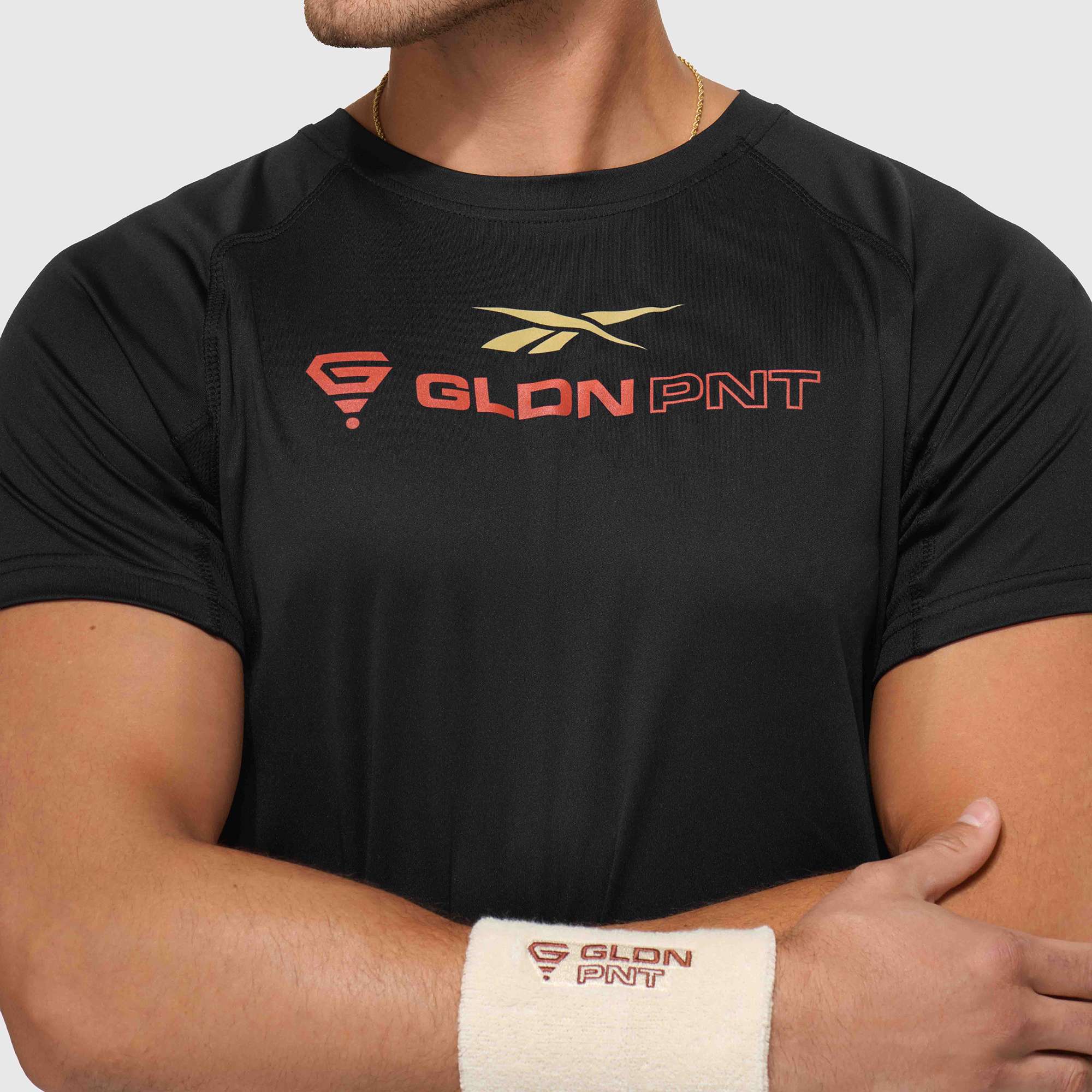 A man crosses his arms wearing the Reebok x GLDN PNT Tech Tee in black with a gold GLDN PNT logo on the chest, paired with a white wristband featuring matching branding. The background is plain light gray.