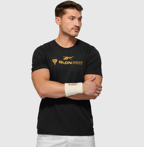 A man wears a black Reebok x GLDN PNT Jersey Tee with yellow lettering and a white wristband, standing with arms crossed and looking to the side against a plain light gray background.