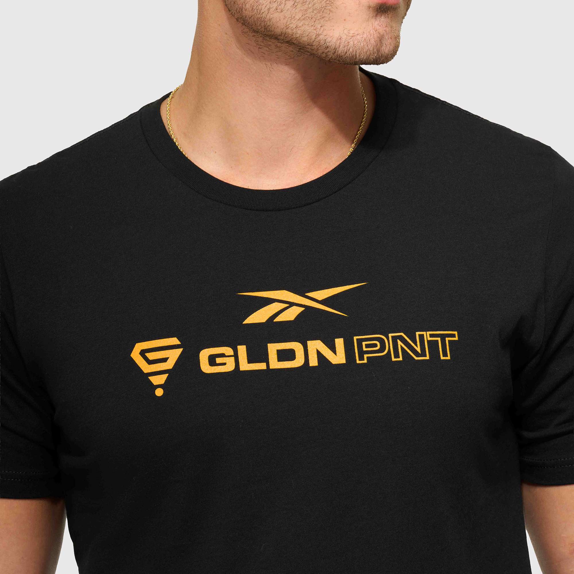 A person wears the Reebok x GLDN PNT Jersey Tee, a minimalist black unisex t-shirt by Reebok with gold graphics and 'GLDN PNT' text, made from premium cotton. Their face is partially visible as they look to the side.