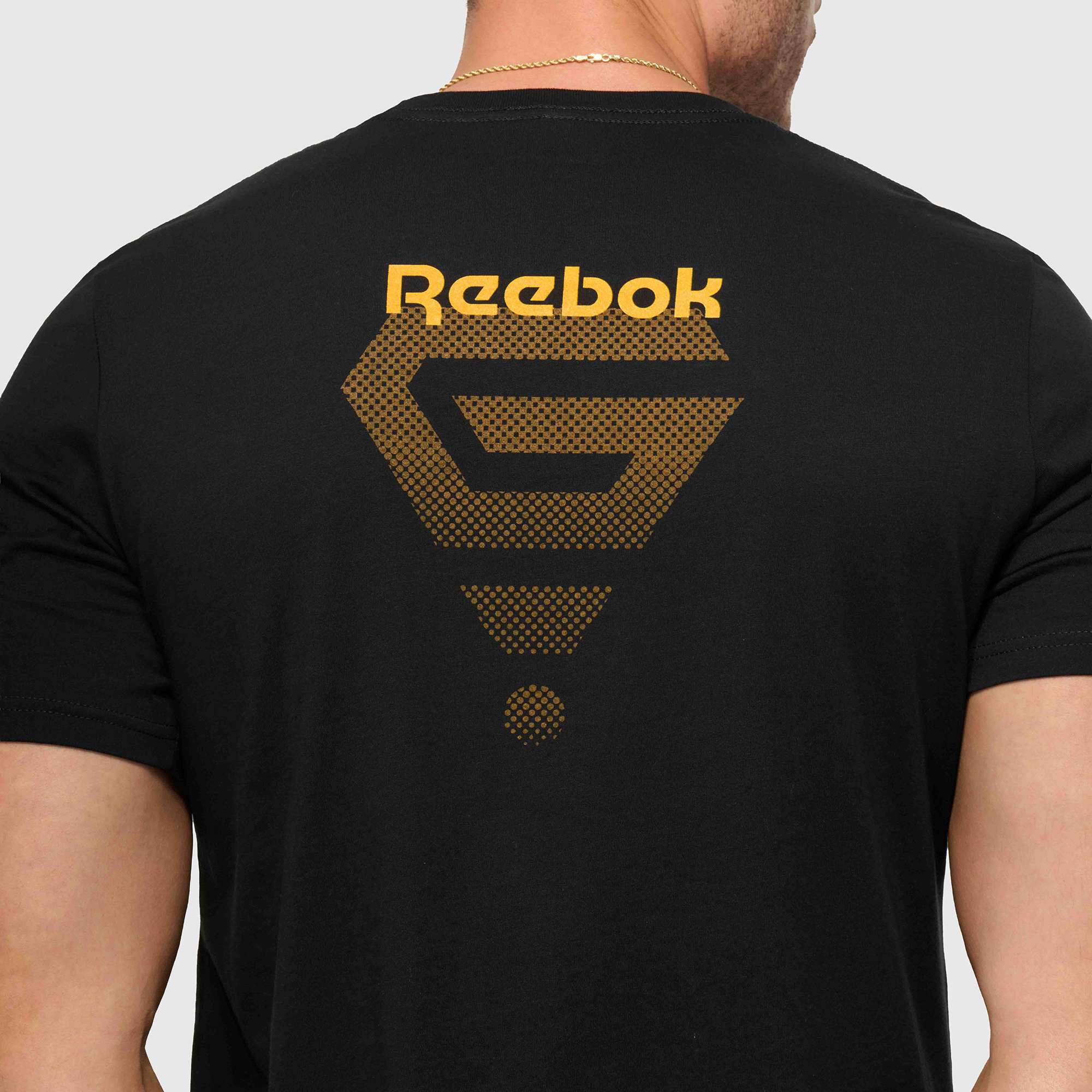 A person wears the Reebok x GLDN PNT Jersey Tee, a premium cotton unisex shirt with a black base, gold Reebok logo, and geometric back design. The image highlights the upper back and neck of this minimalist tee.