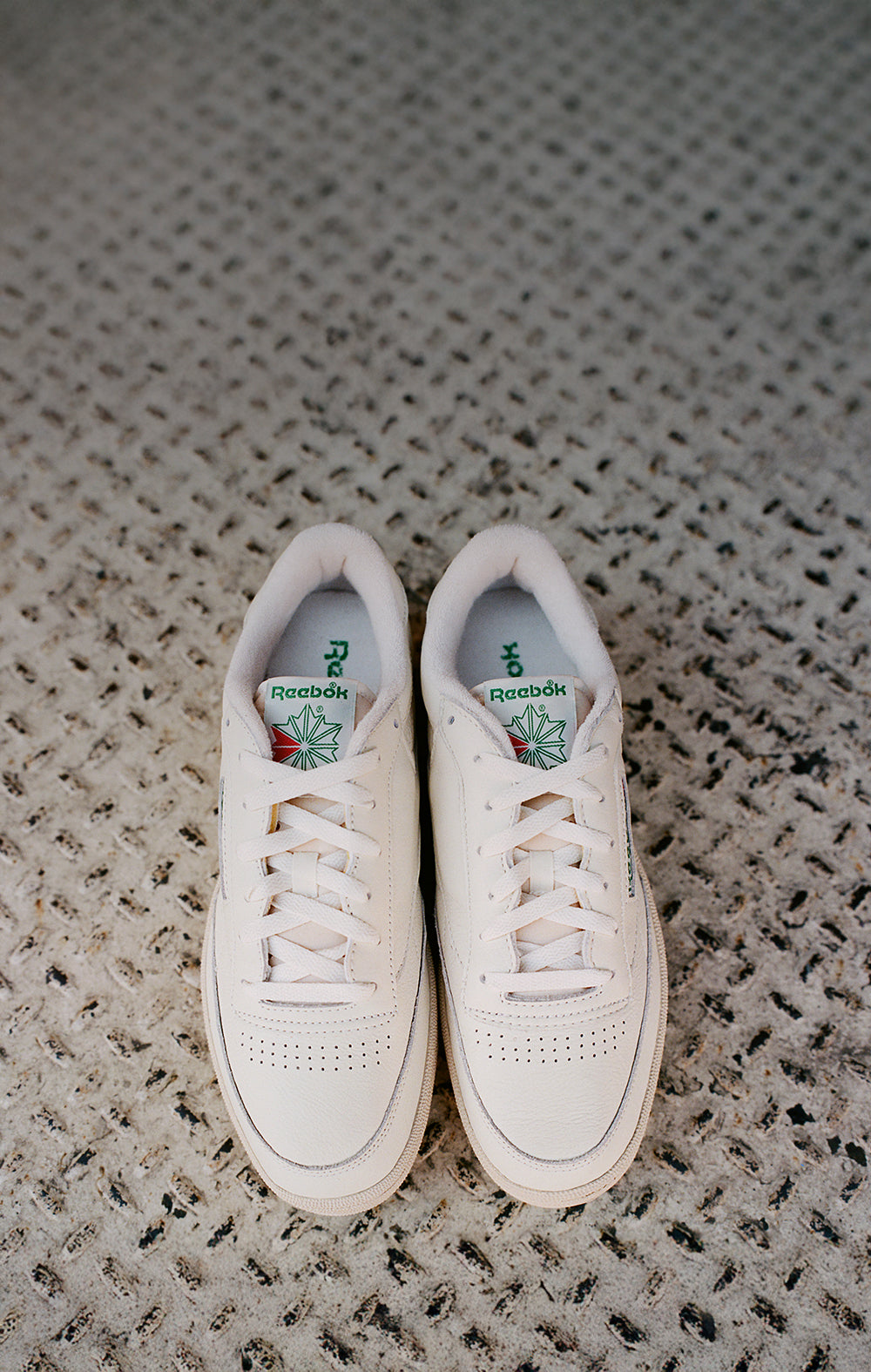 A pair of white Reebok sneakers with green and red logos are placed side by side on a textured, light gray carpet.