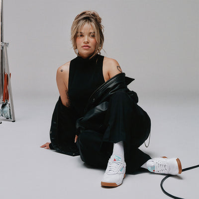 Karol G wearing a black outfit with white sneakers on a plain background
