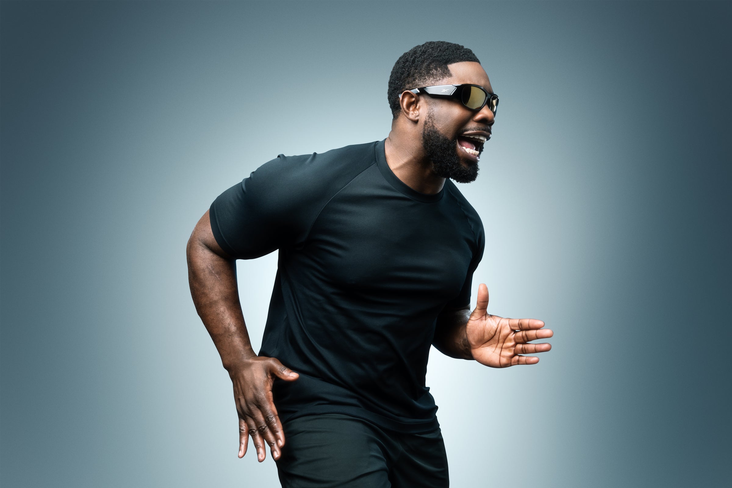 A man in a black athletic outfit and sunglasses poses energetically against a gradient background, appearing to run or shout, with his right arm bent forward and left arm back.