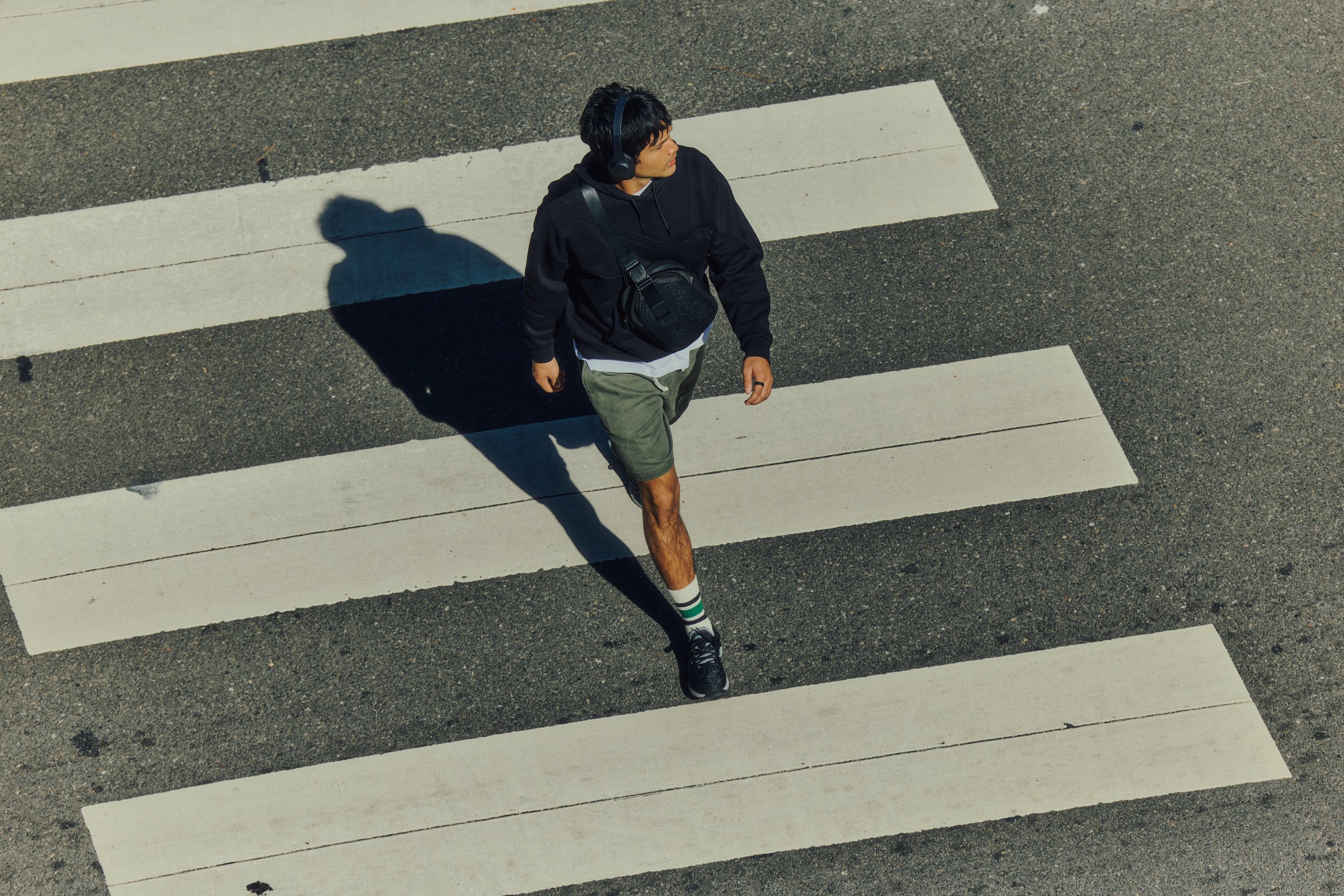 A person wearing headphones, a dark sweatshirt, green shorts, and sneakers walks across a crosswalk on a sunny day, casting a long shadow on the asphalt.