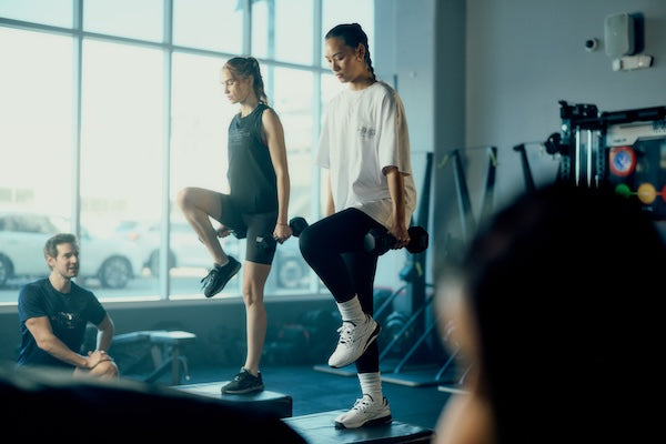 Two women exercise indoors, lifting dumbbells during a step workout, while a man watches and coaches. Large windows in the background let in natural light, and gym equipment is visible.