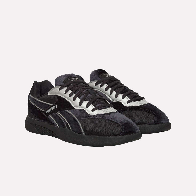 A pair of Pleasures Hammer Street Shoes by Reebok in black and silver, featuring PLEASURES branding on the side and a sleek velvet upper, displayed against a plain white background.