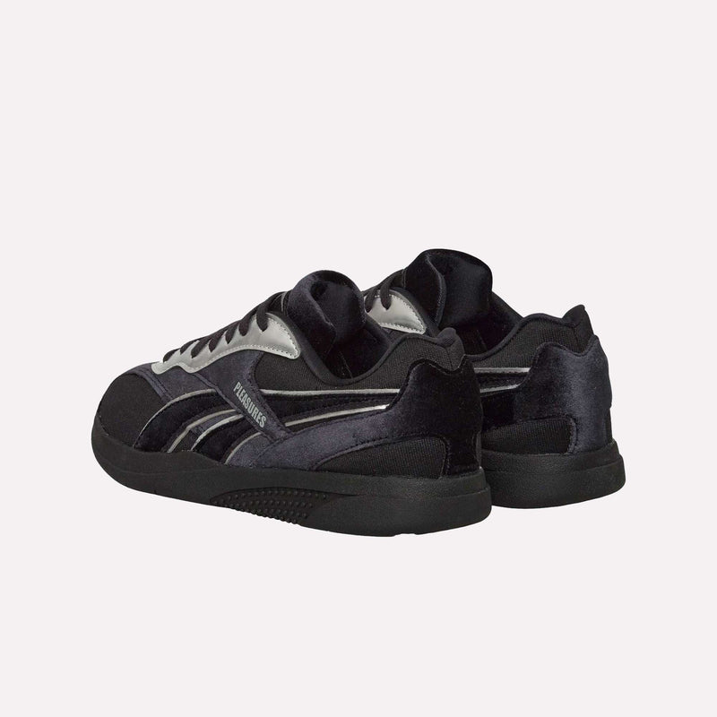 A pair of Pleasures Hammer Street Shoes by Reebok, featuring a black and gray color scheme inspired by classic Reebok indoor soccer shoes, is shown on a white background, positioned at an angle with the heels slightly facing forward.