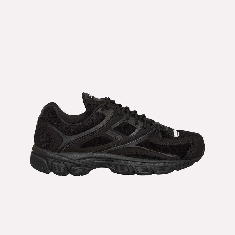 A black athletic sneaker featuring mesh and suede-like panels, black laces, and a chunky black sole, viewed from the side on a plain light background—these standout shoes are the Reebok Pleasures Premier Trinity KFS, a collaboration that merges contemporary style with streetwear influence.