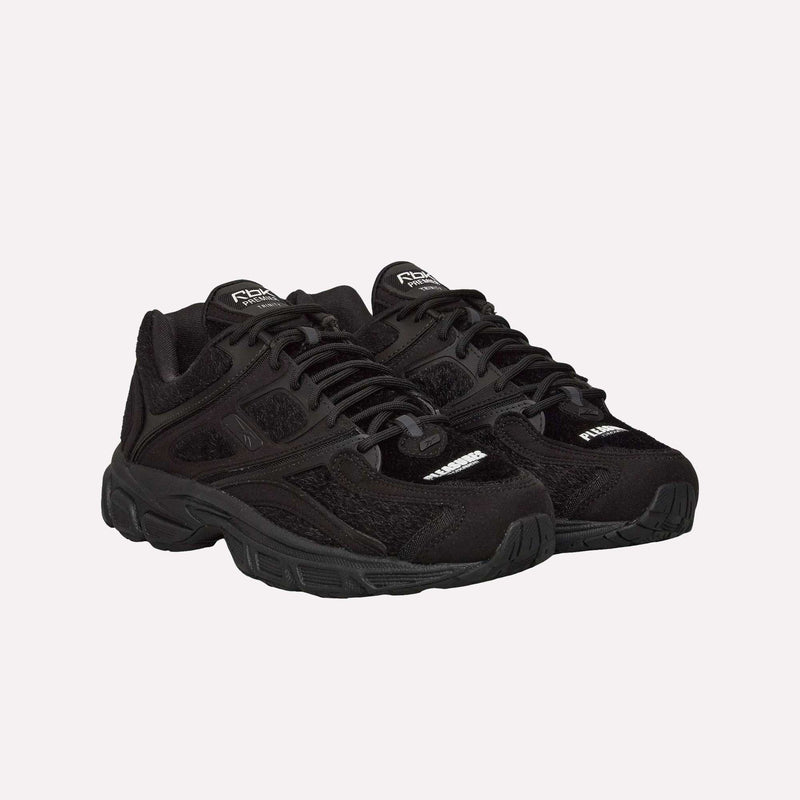 A pair of Pleasures Premier Trinity KFS Shoes by Reebok in black, featuring a pony hair upper, textured detailing, thick soles, and laces, are positioned side by side on a plain white background.