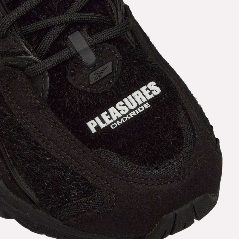 Close-up of the Pleasures Premier Trinity KFS Shoes in black, showcasing textured suede and mesh with “PLEASURES DMX RIDE” in white text on the upper and a small Reebok logo above the laces. As part of the Reebok x Pleasures collaboration, these sneakers blend premium materials with bold branding for a standout look.