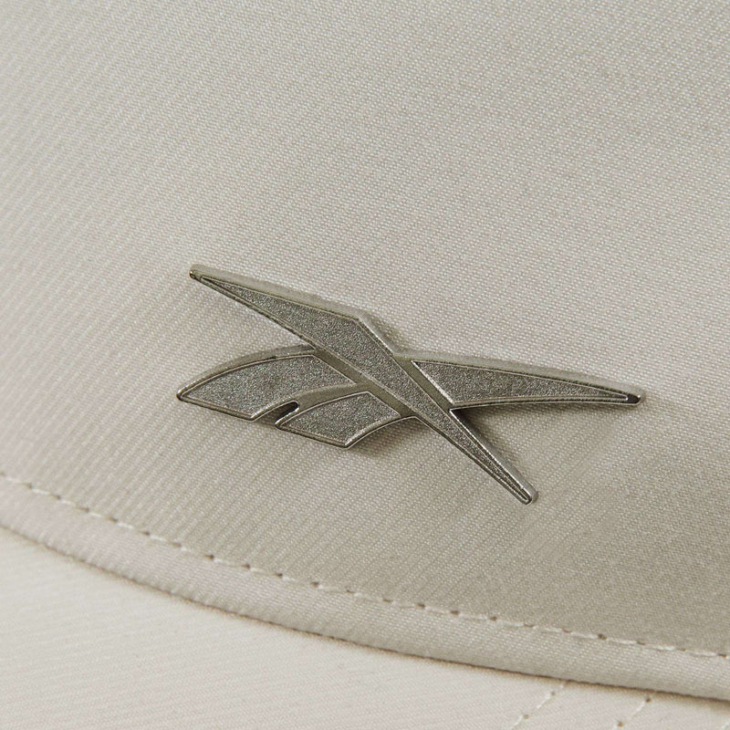 Close-up of the Reebok Badge Cap shows its beige fabric surface with a metallic angular Reebok logo and visible stitching along the edge.