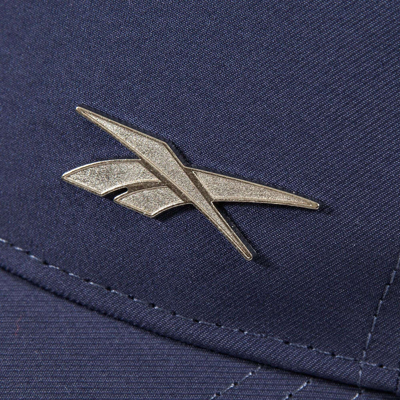 A close-up of a metallic Reebok logo pin on the Badge Cap, featuring dark blue fabric with detailed stitching and a shiny logo.