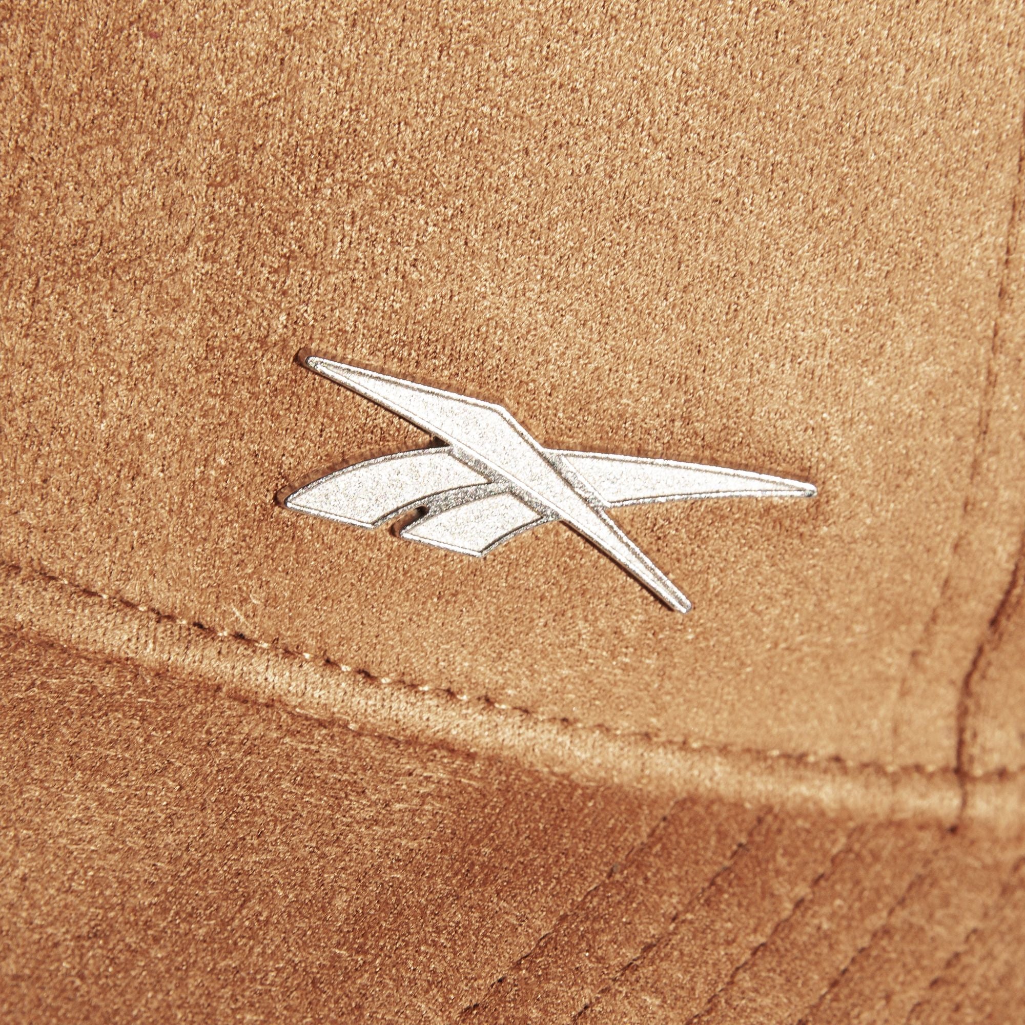 A close-up of the brown Suede Badge Cap by Reebok shows a silver, angular metallic logo, giving this vegan suede cap a modern look.