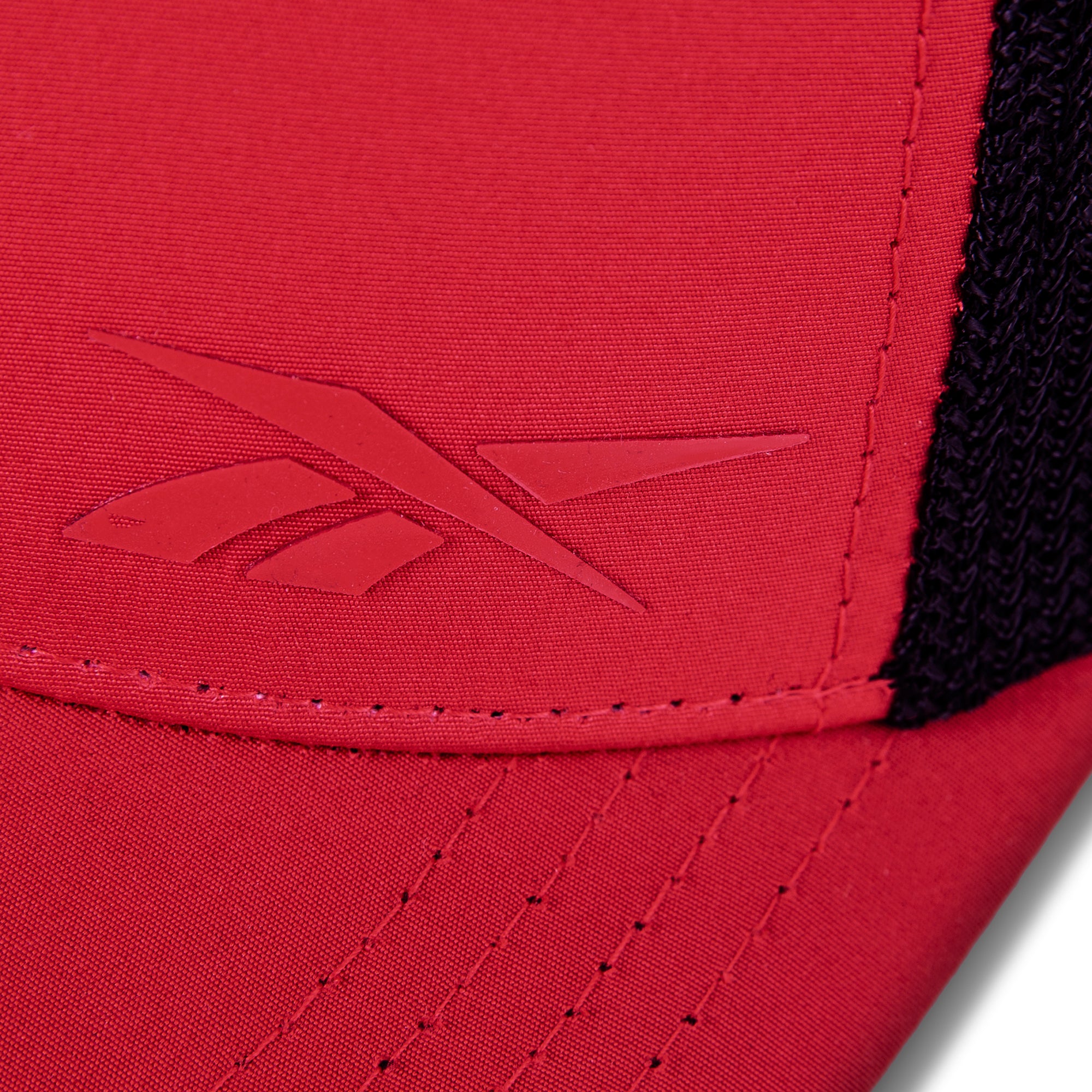 Close-up of the red Reebok Athlete Cap featuring an embossed Reebok logo on the front, black mesh fabric on the side, and detailed stitching along the seam.